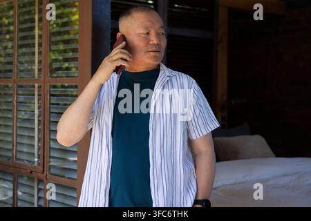 Senior Asian man standing in doorway holding phone to right ear wearing striped shirt, watch, bed Stock Photo