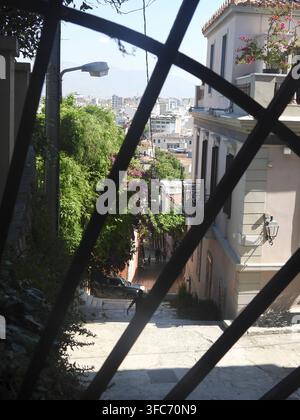 Looking through an antique window grid capturing a sunny view of a narrow, sloping street in Athens with traditional buildings, lush greenery, and dis Stock Photo