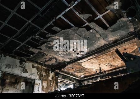 A room interior shows a heavily fire damaged ceiling with an exposed metal grid, missing panels, and visible charring and smoke residue, destruction Stock Photo