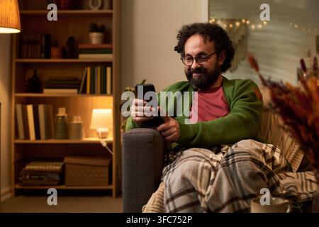 Middle Aged Man Smiling While Using Smartphone on Armchair at Home Stock Photo