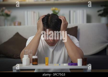 A young Asian man sits at a table and holds his head in pain while looking at various bottles of pills and a glass of water. He seems distressed and u Stock Photo