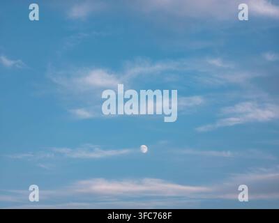 A small full moon appears in a clear blue sky. Light wispy clouds drift past the moon, creating a simple scene during dusk. The setting is calm. Stock Photo