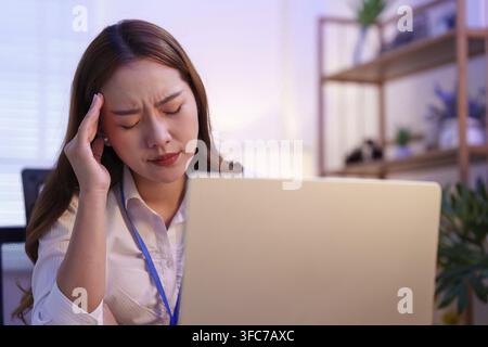 A close-up of a young Asian woman at her desk keeping her head in distress and showing signs of stress and burnout Stock Photo