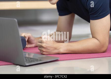 A dedicated young man performs a plank exercise at home using a laptop as a guide. Focus on fitness and online training Stock Photo