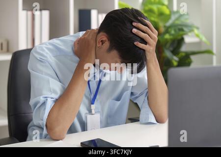 A young Asian man holds his head and rubs his throat. He looks overwhelmed and stressed out on his desk. This image vividly conveys the effects of wor Stock Photo