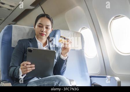 Asian businesswoman makes purchases online using tablet and credit card while traveling on an airplane. She focuses on her digital transaction Stock Photo