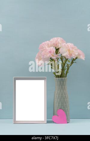 A decorative set with pink carnations in a vase and an empty photo frame on a blue background, symbolizing love and memory Stock Photo