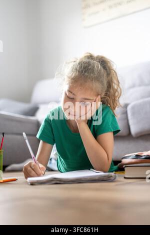 Smiling schoolgirl lying on the floor and writing in a notebook while doing homework at home surrounded by school supplies. Stock Photo