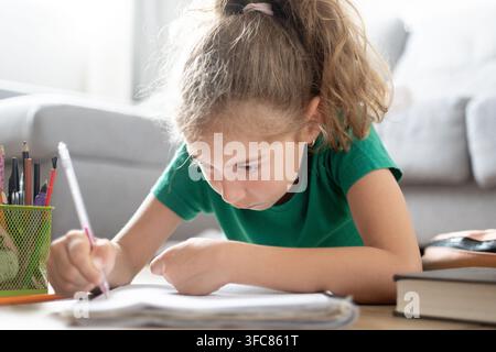 Smiling schoolgirl lying on the floor and writing in a notebook while doing homework at home surrounded by school supplies. Stock Photo