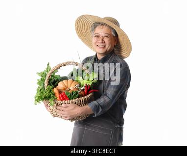 Cheerful senior Korean male farmer in a straw hat and apron holding a basket of fresh vegetables on white. Stock Photo