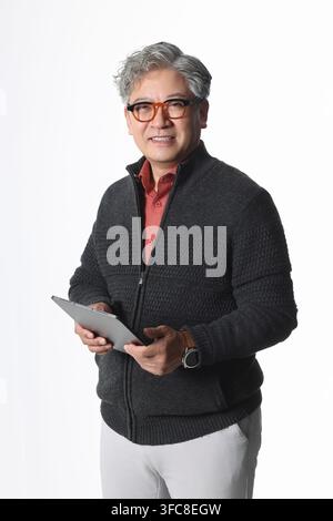 Tech-savvy senior Korean man using a digital tablet in a casual cardigan against a white background. Stock Photo