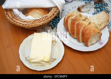 Traditional rustic breakfast table setting with a block of dairy butter on a vintage porcelain plate, sliced crusty white bread, a wicker basket and a Stock Photo