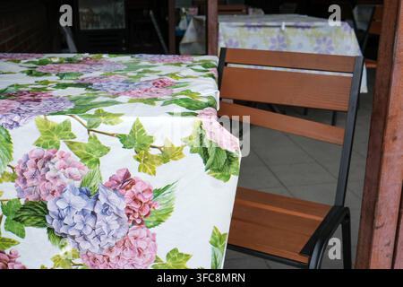 Cozy outdoor cafe table covered with a white tablecloth with a bright pink and purple hydrangea flower pattern. Close up view of a rustic wooden chair Stock Photo