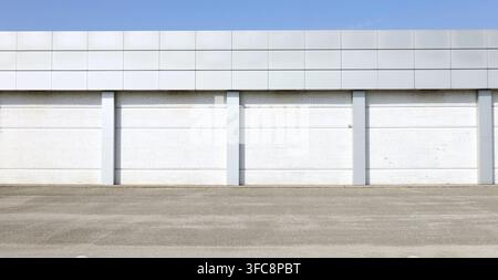 Industrial building made of white brick wall and gray metallic panels on top and columns. Concrete sidewalk and asphalt road in front. Background Stock Photo