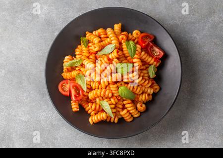 Top view of fusilli pasta with tomato sauce, fresh basil and cherry tomatoes on a black plate Stock Photo