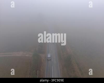 An atmospheric aerial perspective captures a single car navigating a winding country lane through dense fog and mist. Stock Photo