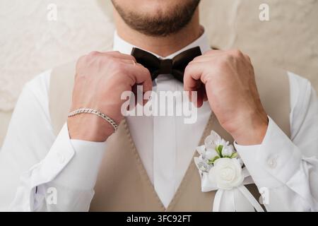 Unrecognizable groom in a white shirt and beige waistcoat with a white rose boutonniere adjusts his black bow tie before the ceremony. Stock Photo