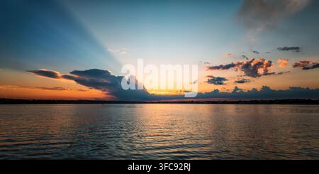 Dramatic sunrays breaking through dark clouds at sunset over calm lake, orange and teal sky, city silhouette on horizon, wide landscape. Stock Photo