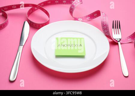Paper note with word Anorexia, plate, cutlery and measuring tape on pink background, closeup Stock Photo