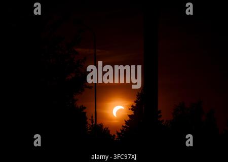 Wide landscape view of a partial solar eclipse low on the horizon behind silhouetted trees and street lamp. Stock Photo