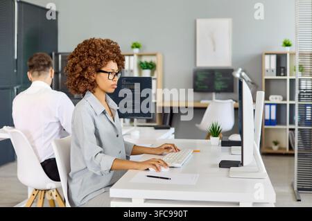Female programmer coding at desktop in modern office with coworker in background Stock Photo