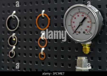 Analog pressure gauge with brass fitting and rubber O-ring seals hanging on dark perforated pegboard wall Stock Photo