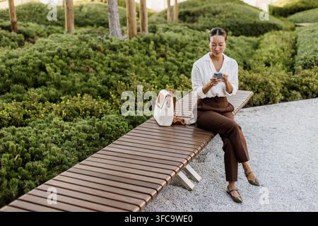 A Korean woman sits on a park bench, joyfully engaging with her smartphone amidst lush greenery. The sun shines, creating a tranquil atmosphere. Stock Photo