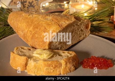 Freshly baked artisan bread with butter and a portion of red caviar are presented on a plate against a warm holiday backdrop of candlelight Stock Photo