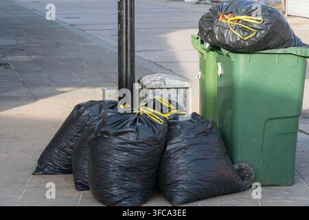 Black garbage bags tied with yellow strings sit piled on a city sidewalk next to an overflowing green wheeled trash bin Stock Photo