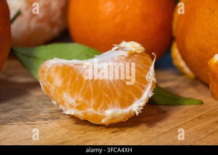 A close-up shot highlights a single peeled segment of a mandarin or clementine resting on a wooden surface with green leaves and whole fruits in the b Stock Photo