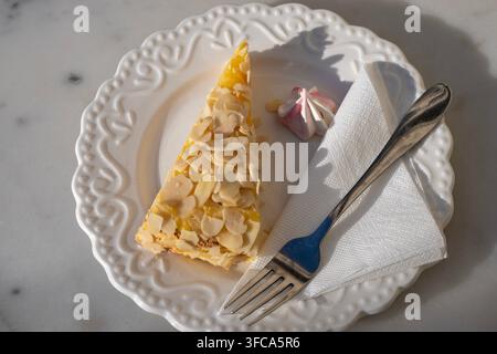 A slice of almond tart topped with flaked almonds is served on an ornate white ceramic plate alongside a small meringue, paper napkin, and fork under Stock Photo