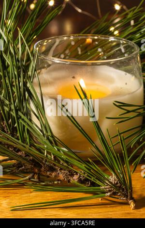 A glowing scented candle in a clear glass jar is nestled among fresh pine branches, Stock Photo