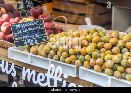 Fresh mirabelle plums lie for sale at a market stall on Rue Braconnot in Nancy, Grand Est, France, with a sign naming the protected designation "Mirabelle de Lorraine." The small yellow plum has held EU Protected Geographical Indication (PGI) status since 1995; Lorraine produces around 90 percent of the world's mirabelle harvest. Peaches are stacked in the background. Stock Photo