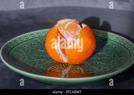 A partially peeled fresh mandarin orange sits centered on an ornate green patterned plate against a dark background. Stock Photo
