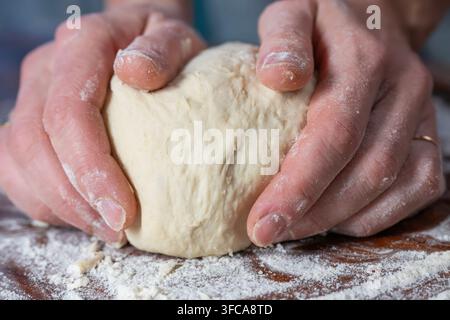A pair of hands kneads a ball of white flour dough on a floured wooden surface Stock Photo