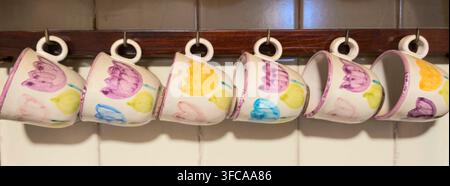 Hand painted ceramic mugs featuring colorful tulip designs, hanging upside down on a wooden rack in a kitchen, representing cozy home decor and drinkw Stock Photo