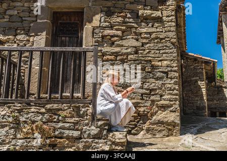 Woman using smartphone while sitting on stone steps in medieval village of Rupit, Catalonia, Spain, checking travel information Stock Photo