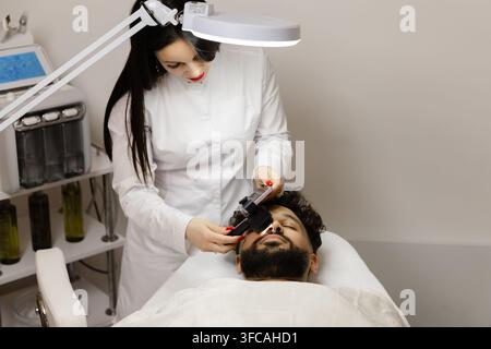 Female doctor using a dermatoscope to examine a male patient's facial skin for dermatology and skincare purposes in a clinic Stock Photo