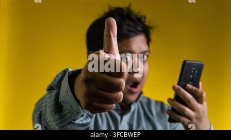 A young Asian male gives a prominent thumbs up gesture towards the camera while holding a modern mobile device, set against a solid yellow background. Stock Photo