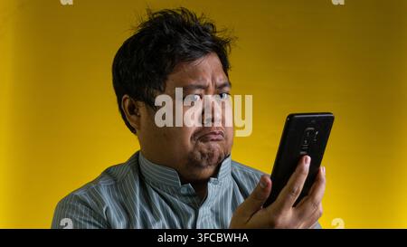 A young Asian male looks at a modern mobile device with a deep frown and a highly disappointed facial expression, standing against a solid yellow back Stock Photo