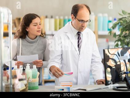 Experienced pharmacist helps girl buy medicine - he reads the prescription and compares it with data on computer Stock Photo