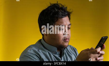 A young Asian male focuses intently on a modern mobile device while standing against a solid yellow background. The individual maintains a serious and Stock Photo