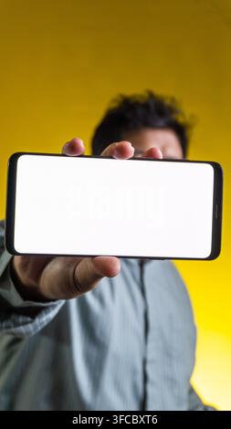 A blurred Asian male presents a smartphone horizontally with a glowing blank white screen directly to the camera against a vivid yellow background, of Stock Photo
