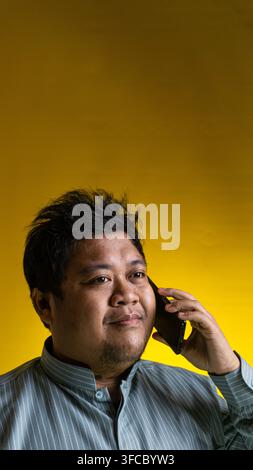 A contented young Asian male engages in a pleasant phone conversation while looking away from the camera against a bright yellow background, radiating Stock Photo