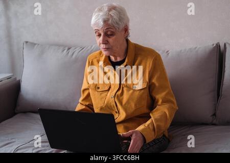 Elderly 70s woman sit on sofa holds laptop feels comfortable using modern tech, surfing website search useful info enjoy personal development. Technology and people concept. Stock Photo