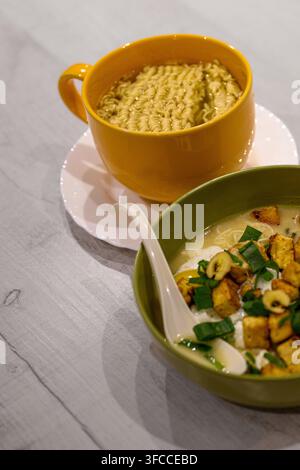 Yellow mug holds instant noodles. Green bowl holds creamy soup with toppings. White spoon rests beside the bowl. Wooden table holds both dishes. Simpl Stock Photo