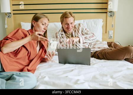 Couple Using Laptop and Smartphone While Relaxing on Bed in Hotel Room Stock Photo