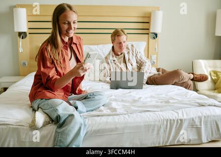 Smiling Woman Using Smartphone with Man Working on Laptop in Hotel Bed Stock Photo