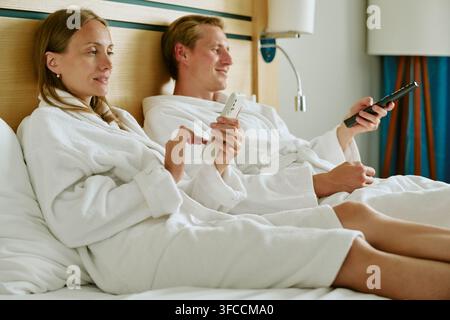 Couple in Bathrobes Relaxing in Hotel Bed Using Smartphone and Remote Stock Photo