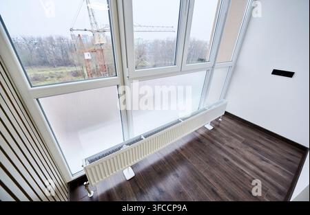 Radiator installed under large window in empty modern room, close up view of heating system and wooden floor. Home interior, energy efficiency and apartment renovation concept Stock Photo